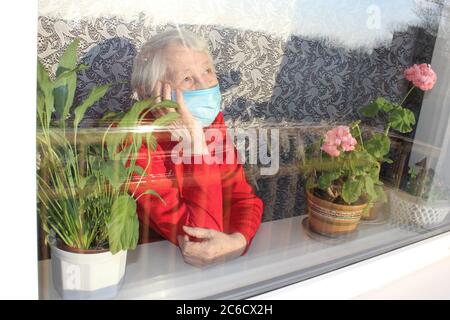 Die Covid-19, Gesundheit, Sicherheit und Pandemie Konzept - ältere alte einsame Frau trägt schützende medizinische Maske sitzt in der Nähe des Fensters zu Hause zum Schutz vor Viren Stockfoto