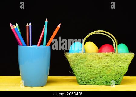 Osterkorb voller bunt bemalter Eier und Becher farbiger Bleistifte, die auf Holztisch stehen, isoliert auf schwarzem Grund. Zusammensetzung mehrerer Farben Stockfoto