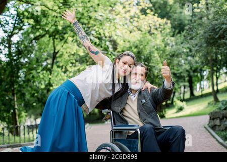 Glückliche junge Frau in blauem Rock und weißem Hemd, mit Dreadlocks, lächelnd, umarmt mit ihrem älteren Großvater im Rollstuhl im Park. Sozialarbeiter Stockfoto