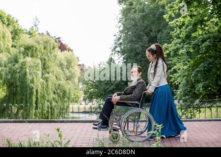 Seitenansicht der jungen modernen Hipster Frau mit langen Dreadlocks, die ihren behinderten Großvater im Rollstuhl im Park unterstützt. Glücklicher älterer Mann Stockfoto