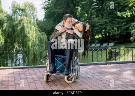 Der alte bärtige Mann im Rollstuhl und seine hübsche Hipster-Enkelin mit Dreadlock-Haaren gehen im Park, umarmen und schauen sich gegenseitig an Stockfoto