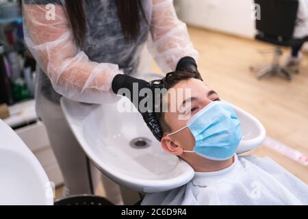 Junger Mann mit einer Schutzmaske, der sich auf einen Hecht stützt, während er sich in einem Friseursalon mit Handschuhen den Kopf wäscht Stockfoto