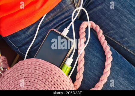 Ein Mädchen sitzt auf einer Bank im Park, während es ein Smartphone von einer externen Powerbank lädt Stockfoto