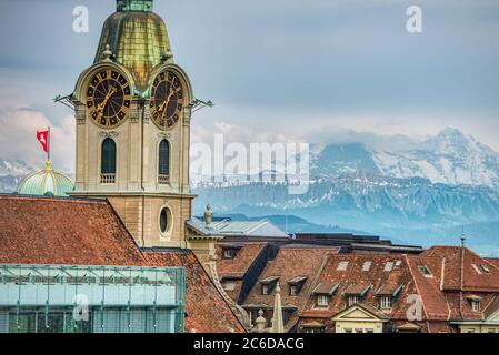 Heiliggeistkirche Glockenturm oder Uhrturm ragt von den gedeckten Dächern der Stadt gegen die verschneiten und majestätischen Alpen - Bern, hervor Stockfoto