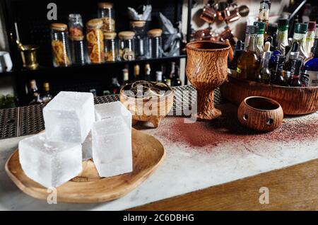 Zutaten für Cocktails in kleinen Flaschen.Schüttler und Zitronenkeile an der Theke. Große Eisstücke auf einem Teller zum Abkühlen von Getränken Stockfoto