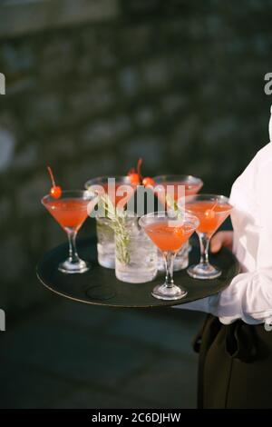 Der Kellner trägt Cocktails auf einem Tablett - Manhattan Cocktail mit Kirschen und Wodka mit Limonade und Rosmarin. Stockfoto