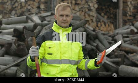 Portrait von Holzfäller Arbeiter posiert in grüner Jacke mit reflektierenden Streifen und hält kleine Axt und Säge auf seinen Händen. Holzfäller stehend und Kamera anblickend. Gesägte Holzstämme, Brennholz Hintergrund Stockfoto