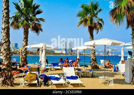 BARCELONA, SPANIEN - AUGUST 19: Badende liegen in Liegestühlen am Strand von Sant Sebastia am 19. August 2014 in Barcelona, Spanien. Im Hintergrund Mapfre Tower Stockfoto