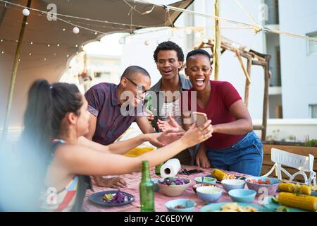 Glückliche junge Freunde, die Smartphone benutzen und am Balkontisch essen Stockfoto