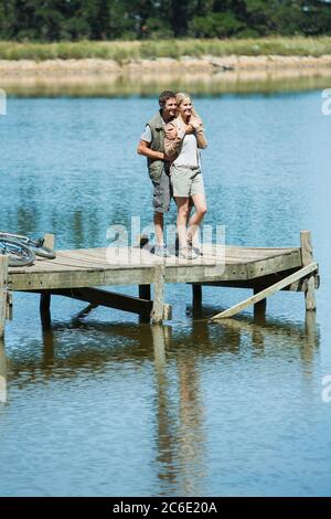 Lächelndes Paar, das sich am Dock über dem See umarmte Stockfoto
