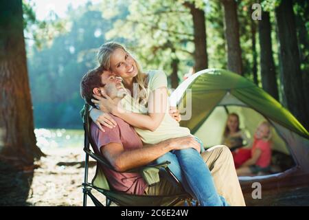 Lächelnde Frau auf dem Schoß des Mannes auf dem Campingplatz im Wald sitzend Stockfoto