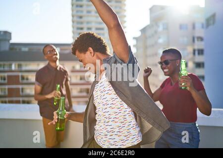 Unbeschwerte junge Freunde tanzen und trinken Bier auf der sonnigen Dachterrasse Stockfoto