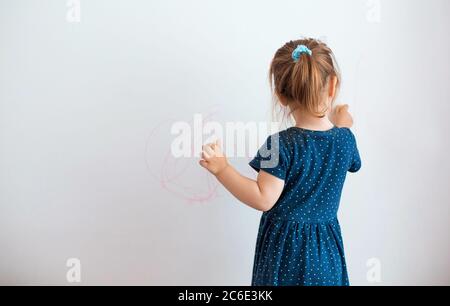 Rückansicht von liebenswert Kind im Kleid stehen in modernen Wohnung und die Schaffung von Bildern auf weißer Wand Stockfoto