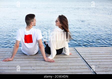 Rückansicht der jungen glücklichen Freundin und Freund sitzen auf hölzernen Kai und einander beim Genießen Wochenende in der Nähe des Sees Stockfoto