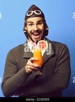 Schöner bärtiger Flieger Mann mit langem Bart auf lächelndem Gesicht hält Glas alkoholischen Cocktail in grau gestrickten Pullover mit Hut und Brille, auf blauem Studio-Hintergrund Stockfoto