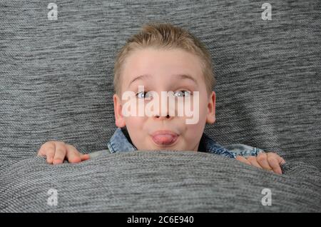 Cute lustige junge ist seine Rücken auf der Couch und macht lustige Gesicht. Spielerischer Junge zeigt Zunge an Kamera, Spaß zu Hause auf dem Sofa. Stockfoto