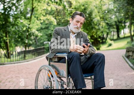 Bärtiger kaukasischer alter Mann sitzt allein im Park im Rollstuhl und nutzt sein Smartphone zum Surfen im Internet oder in den Socail-Netzwerken Stockfoto