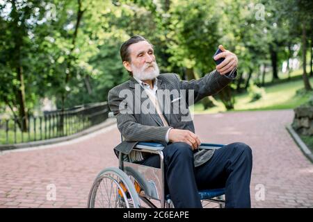 Glücklicher älterer behinderter oder behinderter bärtiger Mann, der auf einem Rollstuhl im Park sitzt, sein Smartphone benutzt, um sich auf dem Hintergrund von zu fotografieren