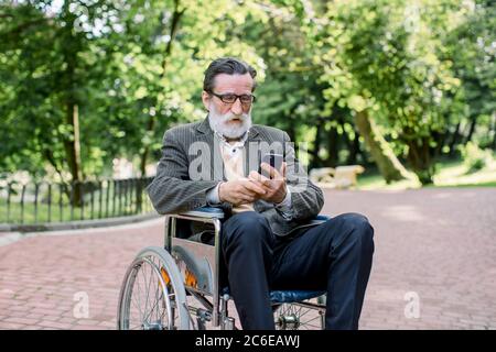 Gutaussehender älterer gebärtiger Mann, der im Rollstuhl im Park sitzt, sich den Kopf beugt, während er auf den Bildschirm seines Gadgets schaut und nach Bedarf sucht Stockfoto