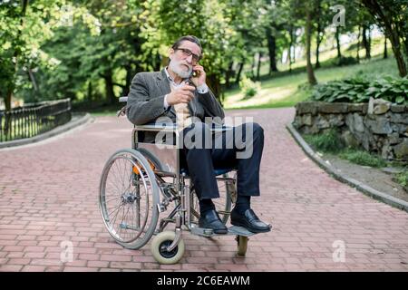 Ein gutaussehender älterer bärtiger Mann im Rollstuhl sitzt mitten in der Gasse im Sommerpark, telefoniert mit jemandem und lächelt Stockfoto