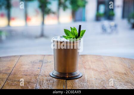 In der Mitte des Bildes sieht man einen Cocktail, der in einer Metalltasse mit Eis und Stroh serviert wird.der Becher ist auf einem Holztisch und dem Hintergrund Stockfoto