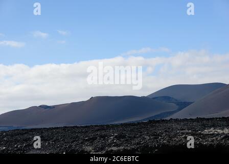 Die weite Leere und Einsamkeit der Lanzarote schwarz gefrorenen Lava vulkanischen Wüste und die vulkanischen Kalderas gegen blau bewölkt im Hintergrund. Stockfoto