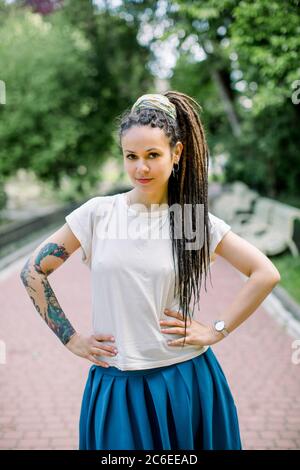 Nahaufnahme Porträt von attraktiven Hipster Mädchen mit Dreadlocks und Tattoos, trägt weißes Hemd und blauen Rock, posiert vor der Kamera in einem Stadtpark in der Stockfoto