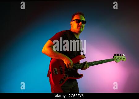 Junge kaukasische inspirierte und expressive Musiker, Gitarrist auf gradienten farbigen Hintergrund in Neonlicht. Konzept der Musik, Hobby, Festival, Kunst. Fröhliche Künstler, bunt, helles Porträt. Stockfoto