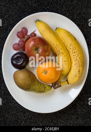 Fruit stil life ariel Blick Bananen Birne orange Pflaume Apfel und einige rote Trauben auf einem weißen ovalen Schale auf einem schwarzen Teppich backgound gesetzt angeordnet Stockfoto