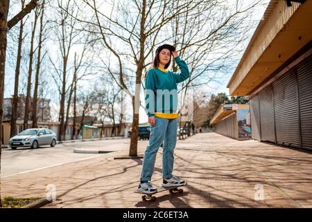Eine junge kaukasische Hipster Frau stellt sich auf einem Skateboard. Im Hintergrund eine Gasse. Konzept des sportlichen Lebensstils und der Straßenkultur. Stockfoto