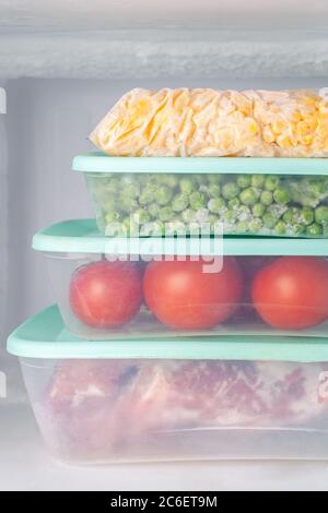 Tiefgefrorenes Gemüse und Fleisch in blauen Kunststoffbehältern. Gefrorener gelber Mais, grüne Erbsen, rote Tomaten, Fleisch. Tiefkühlschrank. Stockfoto