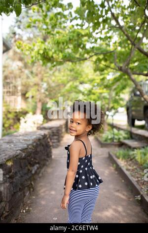 Junge schöne afroamerikanische Mädchen in städtischen Umgebung stehen, während der Blick zurück in die Kamera. Der Hintergrund umfasst einen Gehweg und Bäume. Stockfoto