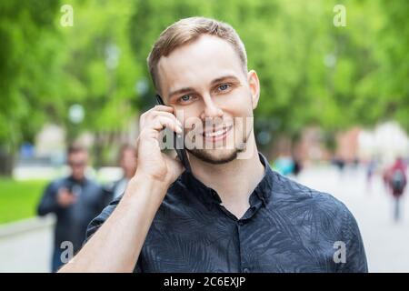 Porträt eines hübschen jungen Mannes, der im Park spazierengeht und auf dem Handy spricht. Mann, der auf dem Handy spricht, Nahaufnahme. Stockfoto