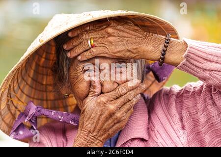 Alte Vietnamesin in violettem Pullover legt ihre Hände auf ihr Gesicht, um ihren Mund und ihre Stirn zu bedecken. Stockfoto