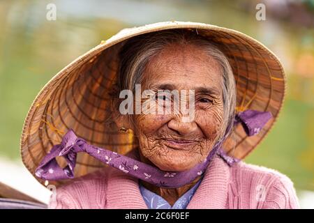 Vietnamesische alte Frau in violettem Pullover und Hut lächelt vor der Kamera Stockfoto