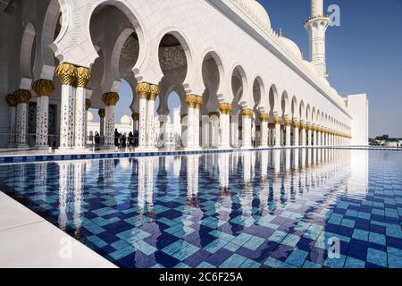 Blick auf die weißen Säulen der Abu Dhabi Moschee, die sich an einem sonnigen Tag mit blauem Himmel im Wasser des Pools spiegeln Stockfoto