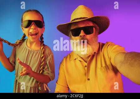 Retro-Mode ist zurück. Älterer Mann verbringt glückliche Zeit mit Enkelin in Neon. Fröhliche ältere Lebensstil, Familie, Kindheit, Tech-Konzept. Mit altmodisch gealtertem Hut und Sonnenbrille. Copyspace. Stockfoto