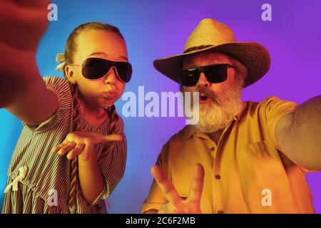 Retro-Mode ist zurück. Älterer Mann verbringt glückliche Zeit mit Enkelin in Neon. Fröhliche ältere Lebensstil, Familie, Kindheit, Tech-Konzept. Mit altmodisch gealtertem Hut und Sonnenbrille. Copyspace. Stockfoto