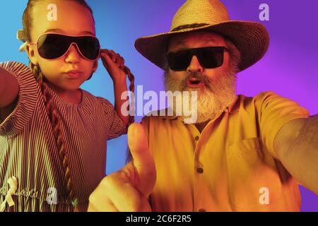 Retro-Mode ist zurück. Älterer Mann verbringt glückliche Zeit mit Enkelin in Neon. Fröhliche ältere Lebensstil, Familie, Kindheit, Tech-Konzept. Mit altmodisch gealtertem Hut und Sonnenbrille. Copyspace. Stockfoto