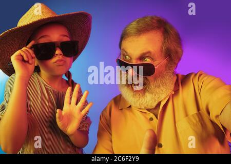 Retro-Mode ist zurück. Älterer Mann verbringt glückliche Zeit mit Enkelin in Neon. Fröhliche ältere Lebensstil, Familie, Kindheit, Tech-Konzept. Mit altmodisch gealtertem Hut und Sonnenbrille. Copyspace. Stockfoto