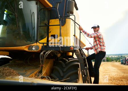 Harvester-Maschinenfahrer klettert in ein Fahrerhaus, um sein Weizenfeld zu ernten. Bauer, der in Mähdrescher auf Leiter hält Geländer. Agronom betrachtet Stockfoto