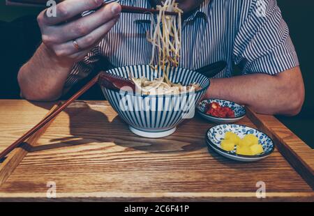 Nahaufnahme des Mannes, der Sojabohnensprossen an Essstäbchen hält und seinen koreanischen Ramen mit Kimchee und Daikon-Rettich-Beilagen isst Stockfoto