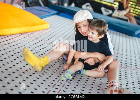 Zwei kaukasische Brüder Jungen genießen es, auf einem Trampolin in einer Umarmung zu sitzen. Kinder spielen zusammen auf einem Trampolin in einem Sportzentrum. Fitnessraum im Freien Stockfoto