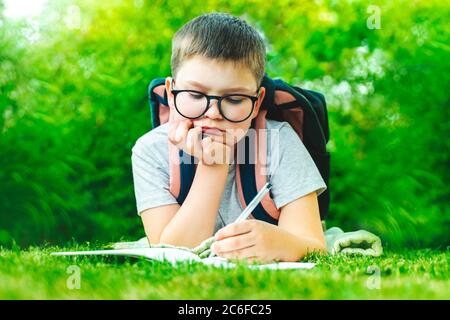 Nette Schuljunge mit Rucksack in Brille auf Gras liegend macht Hausaufgaben auf Gras in der Natur sitzen. Männliche Kind Zeichnung schreiben Notizen tun Mathematik im Freien Stockfoto