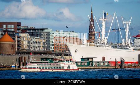 Ein Ausflugsboot vor einem großen Frachtschiff und Bummeltouristen im Hintergrund an den landungsbrücken im Hamburger Hafen mit Kirche Stockfoto