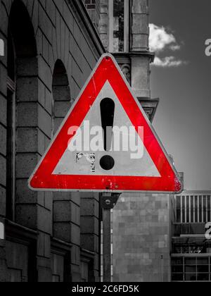 Dreieckiges, rot gerahmtes Straßenschild mit Ausrufezeichen warnt vor einem Gefahrenpunkt neben Gebäuden. Schwarz-Weiß-Fotografie Stockfoto