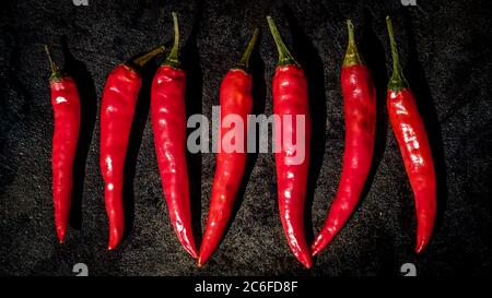 Draufsicht auf eine Gruppe von 7 roten, zerknitterten Chilischoten, die nebeneinander auf einer gusseisernen Oberfläche ausgekleidet sind Stockfoto