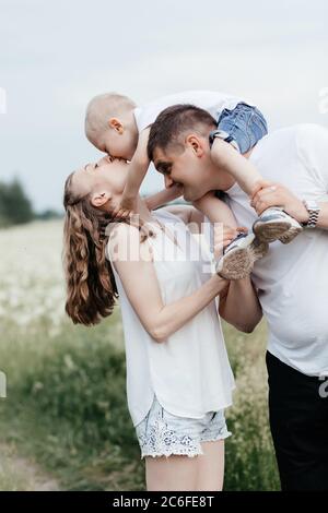 Kleiner Junge, der auf den Schultern seines Vaters sitzt und in diesem Moment Mutter küsst. Junge glückliche Familie. Mutter, Vater und kleiner Sohn Stockfoto