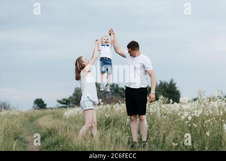 Mutter und Vater heben ihren kleinen Sohn hoch und halten seine Hände, während sie auf der Natur spazieren. Junge glückliche Familie. Mutter, Vater und kleiner Sohn Stockfoto