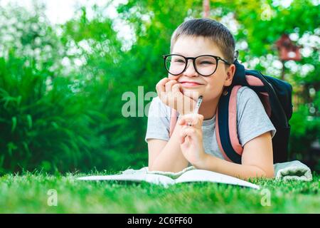 Glücklich Schuljunge in Brille Studium im grünen Park Blick auf Kamera schreiben im Buch Hausaufgaben. Lächelndes Kind macht Notizen im Freien im Park. Zurück t Stockfoto